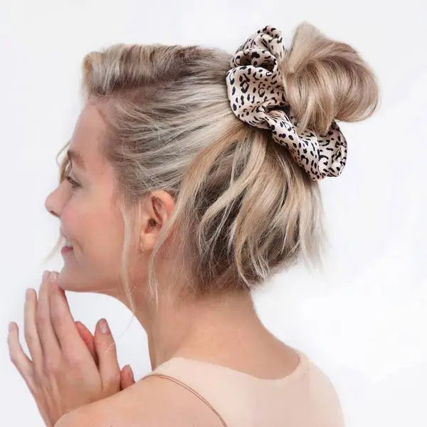 A woman with blonde hair styled in a loose bun using a large, ECO FRIENDLY BRUNCH SCRUNCHIE by KITSCH, is smiling and looking to the side with both hands near her face. She is wearing a light, sleeveless top against a white background.