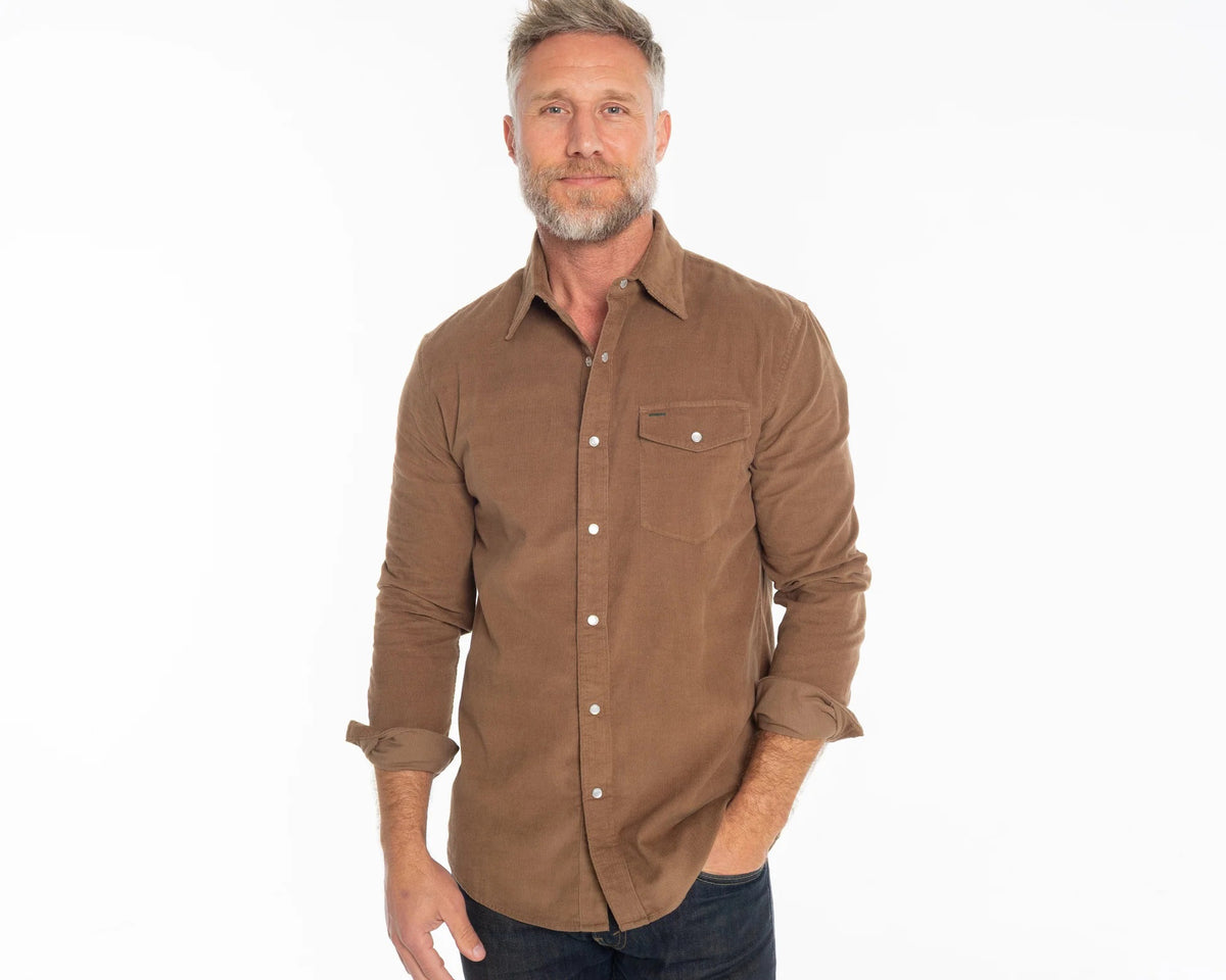 A man with short gray hair and a beard stands against a white background, wearing the CRIQUET Corduroy Pearl Snap Shirt in Walnut with rolled-up sleeves and dark jeans, smiling slightly with one hand in his pocket.