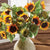 A bouquet of vibrant NAPA HOME AND GARDEN SUNFLOWER STEMs, with their crafted green leaves, is arranged in a beige ceramic vase. The sunflowers, featuring bright yellow petals and dark brown centers, enhance the home decor as they are displayed indoors near a window with a wooden background.