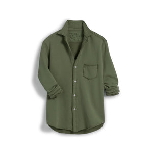 In a sunny, rustic kitchen, a woman smiles while wearing the FRANK & EILEEN EILEEN Relaxed Button Up in Triple Fleece Caper, paired with black wide-leg pants and sandals. She touches her hair as sunlight fills the cozy space.