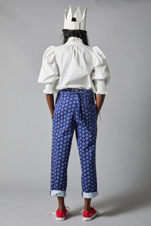 Rear view of a person wearing the Hope for Flowers Smocked Shirt, a bright red blouse with puffed sleeves, paired with blue jeans featuring pom-poms. Made from GOTS Certified organic cotton, this outfit embodies sustainable fashion against a gray background.