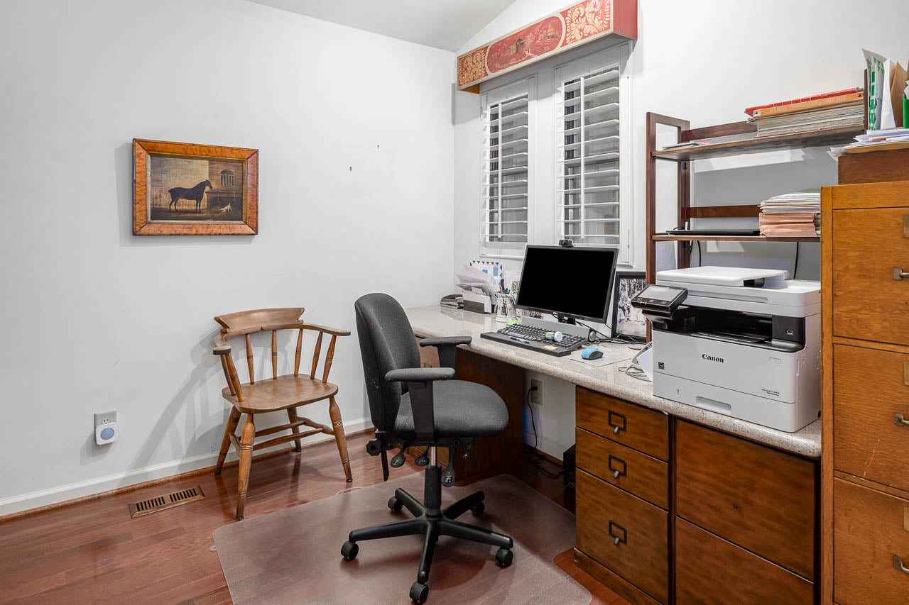 A home office with a desk, computer, printer, office chair, and wooden chair. There are shelves with papers, a filing cabinet, and a framed picture of a horse on the wall. White shutters cover the window above the desk.