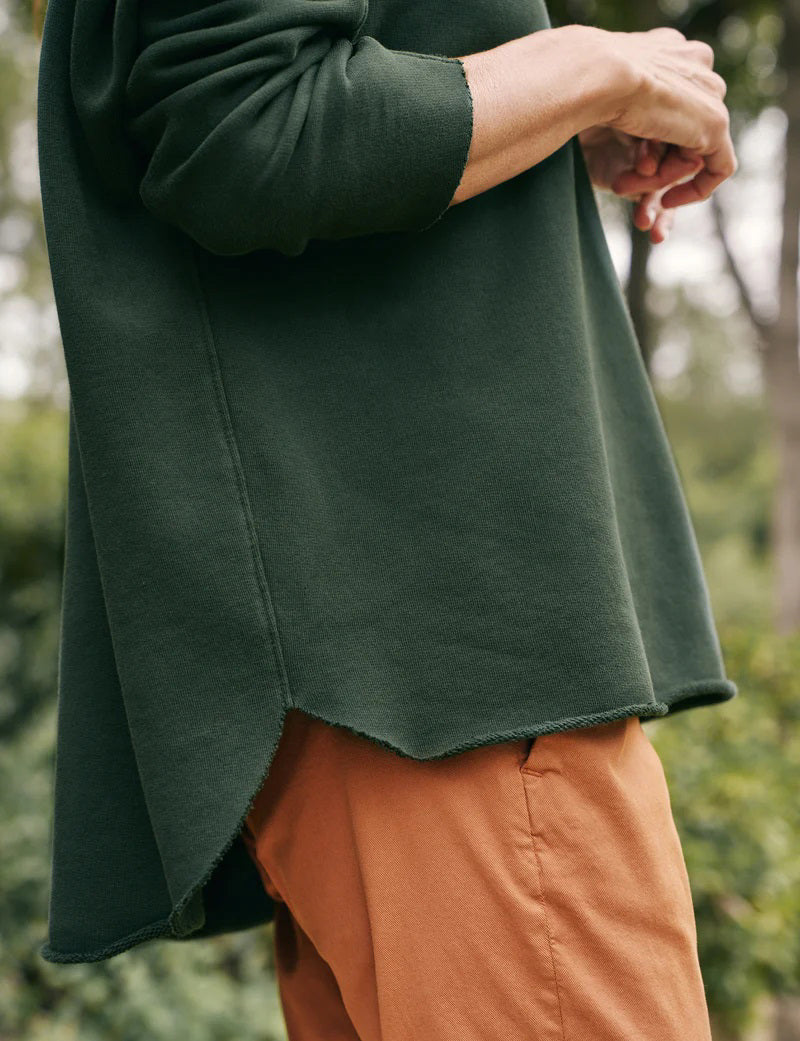 Close-up image of a person standing outdoors, wearing the FRANK & EILEEN Anna Long Sleeve Capelet in Evergreen with cozy sleeves and orange pants. Only part of their upper body and arms are visible. The scene is set against a blurred background of greenery.