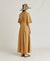 A woman stands in profile wearing the MOMONI - MARTINELLA DRESS, a long, tan cotton poplin dress from MOMONI with short sleeves and a flared skirt. She pairs it with a wide-brimmed, patterned hat, brown sandals, and a necklace. She faces left against a plain, light background.