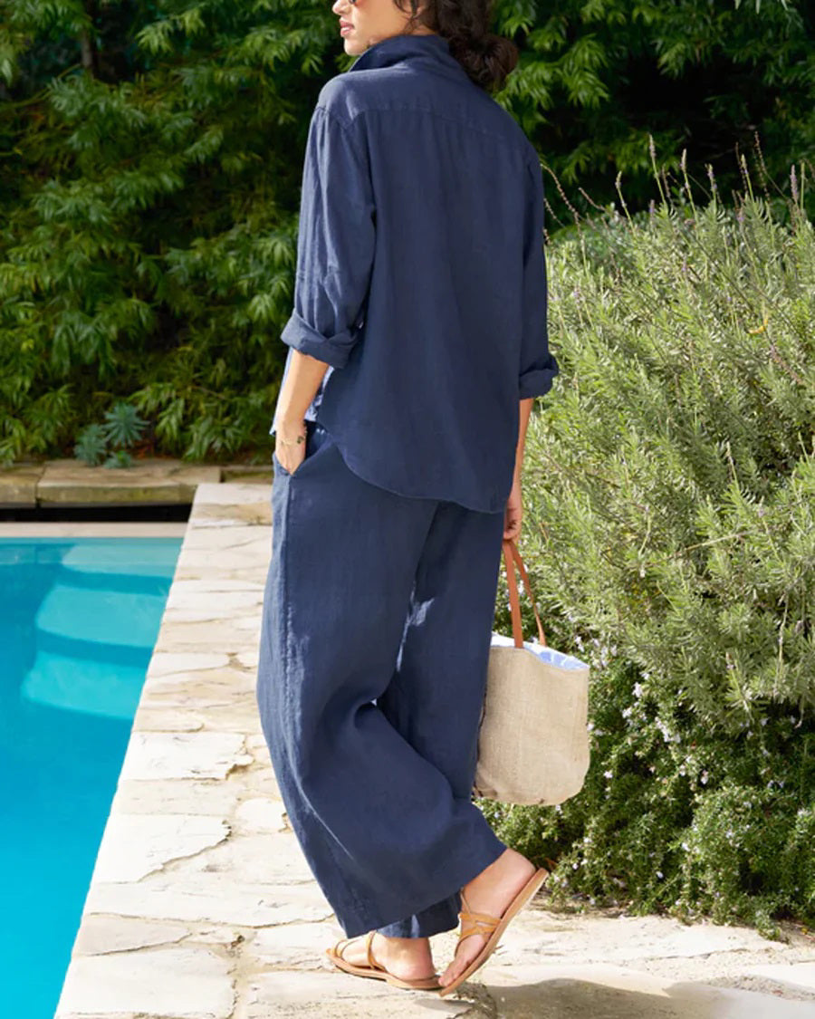 A person wearing a relaxed, navy blue button-up shirt and matching wide-leg silhouette pants from the FRANK & EILEEN Tuscany Getaway Set, including the FRANK AND EILEEN - MAISIE WIDE LEG LINEN PULL ON PANT, with hands in pockets, stands poolside on a sunny day. They carry a beige woven tote bag with blue and red stripes, have short curly hair, and wear sunglasses and sandals.