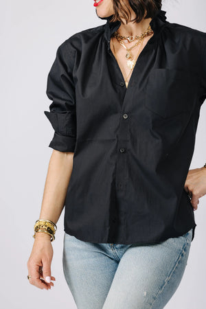 A woman is pictured from the shoulders down, wearing the versatile KMJ - QUARANTINA SHIRT, a white button-up with rolled-up sleeves, paired with blue jeans. She has her hands in her pockets and accessorizes with layered gold necklaces and bracelets. Her nails are manicured and painted white.