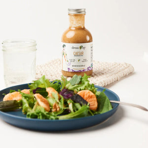 A 10 fl oz (295 ml) bottle of Dress It Up Dressing - Caesar dressing. The label showcases an illustration of a person in a green dress and emphasizes the product's low-calorie formulation made with olive oil and chia seeds, highlighting its simple, wholesome ingredients. The bottle is topped with a metal screw cap.