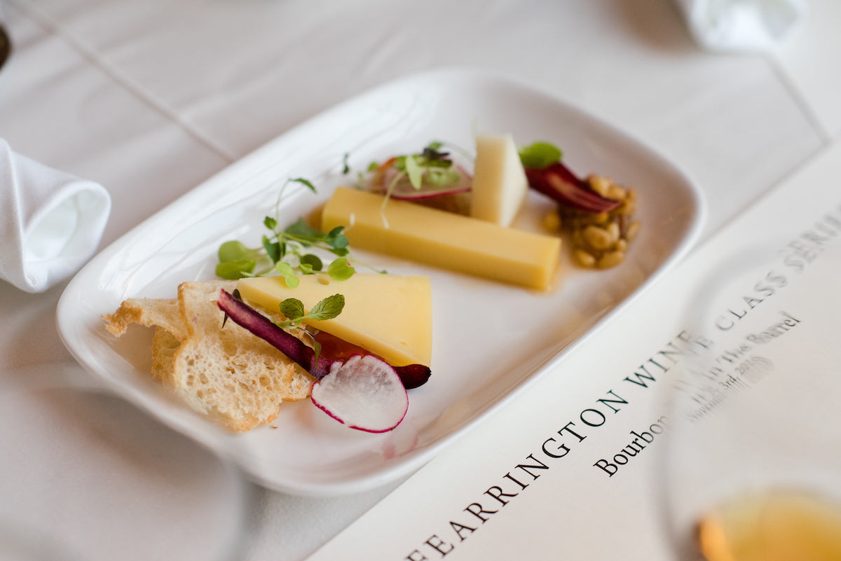 A rectangular white plate with two slices of cheese, microgreens, a slice of radish, a piece of crisp bread, a small dollop of mustard, and a piece of cured meat, set on a white tablecloth next to a wine class seminar menu.