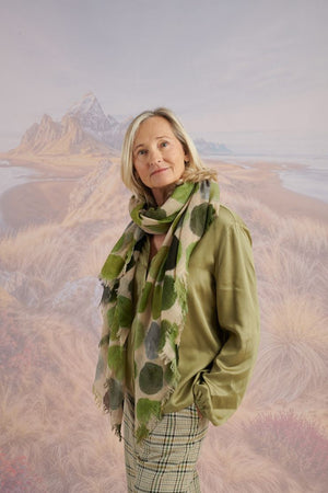 A woman with blonde hair displays the STORIATIPIC - BETTY WOOL SCARF, a large rectangular wool accessory with earth-toned polka dots, set against a scenic mountain landscape.