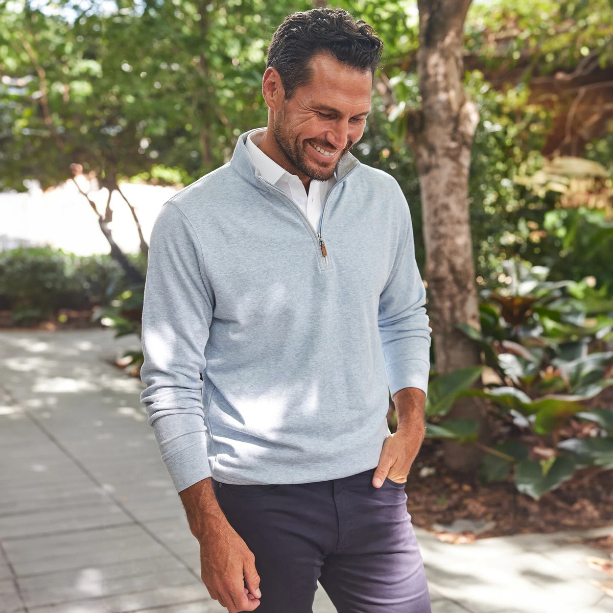 A man smiles outdoors on a sunny day, wearing the COLLARS AND CO Cloud Texture Quarter Zip Pullover in Light Blue over a white polo shirt, with greenery and a shaded path behind him.
