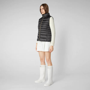 A woman with long brown hair is wearing a white sweater, black shorts, white socks, and black loafers. She is also sporting oversized square glasses and the SAVE THE DUCK - LYNN PUFFER VEST from SAVE THE DUCK made from quilted technical fabric. She stands against a plain gray background.