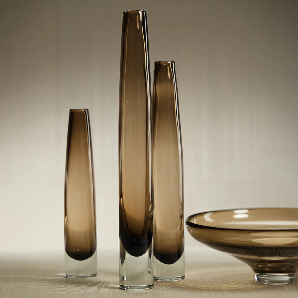 The ZODAX TATE Slim Glass Taupe Vase - Medium features translucent brown glass, a smooth surface, and a clear base. Measuring 2.5 in x 17 in, it is displayed against a plain white background.