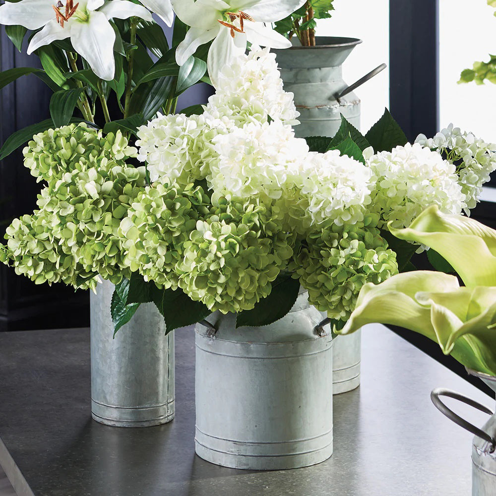 NAPA HOME AND GARDEN cone hydrangea stems are elegantly arranged with large leaves in rustic metal containers on a gray countertop. Lilies and other flowers add a fresh, realistic touch to the display, perfect for any home or office setting.