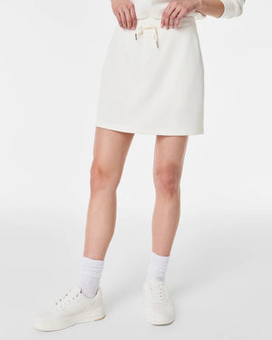 A person stands against a plain white background wearing a matching white long-sleeved sweater and the SPANX - AIRESSENTIALS SKORT by SPANX. Their hair is tied back in a low bun, and they are looking slightly to the side. They complete their look with white ankle socks and sneakers, highlighting the skort's hidden pocket feature perfectly.