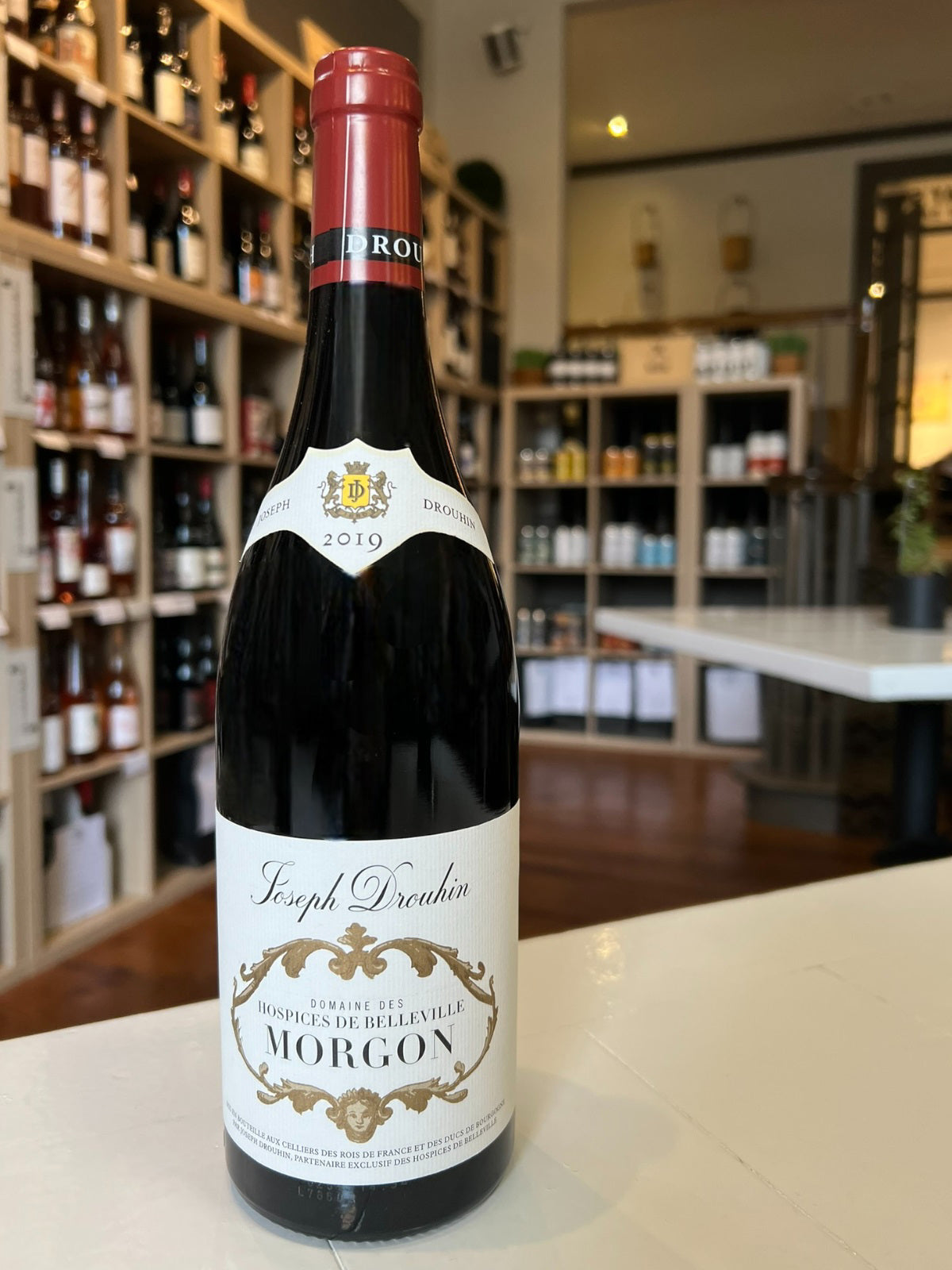 A 2019 bottle of DROUHIN HOSPICE DE BELLEVILLE MORGON BEAUJOLAIS from WINE: GRAPEVINE, made with Gamay grapes. The cream label with gold accents reads &quot;Hospices de Belleville Morgon.&quot; Ideal for Burgundy wine lovers.