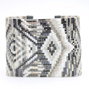 The JULIE ROFMAN - HORIZON CUFF from JULIE ROFMAN JEWELRY is a handwoven glass beaded delica bracelet that showcases an intricate geometric pattern in black, white, and gray. The design includes diamond shapes and symmetrical lines for a mosaic-like appearance, finished with an adjustable buckle clasp to ensure a perfect fit.