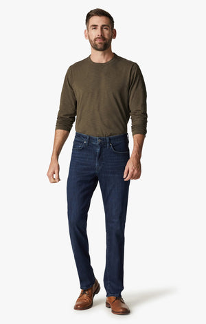 A man in a green long-sleeve shirt, brown shoes, and 34 HERITAGE Charisma Relaxed Straight Leg Jean in Dark Midnight Brushed Urban wash stands with his back to the camera against a plain white background.