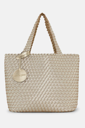 The WOVEN REVERSIBLE TOTE BAG by LINES OF DENMARK is a white tote featuring two shoulder straps and a circular faux-leather tag on one strap. This shopper showcases a textured, interlaced pattern and reversible design, providing stylish and elegant dual styling options. The image background is plain white.