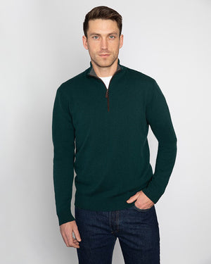 A person is wearing the KINROSS CASHMERE Men's Cashmere Suede Trim Quarter Zip Mock in blue over a white shirt and dark jeans. They have one hand in their pocket and stand against a plain, light-colored background. The quarter-zip sweater features a brown zipper pull.