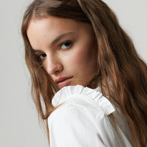 A young woman with long, wavy brown hair stands against a plain gray background. She is wearing the LANHTROPY - JANE COTTON POPLIN SHIRT from LANHTROPY, a white long-sleeved blouse made of 100% cotton poplin, featuring ruffled details at the neck and cuffs, paired with black high-waisted shorts. Her hands are in her pockets, and she has a neutral expression.