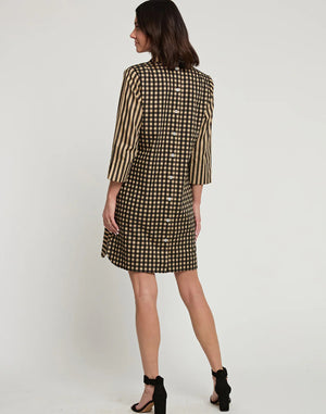 A woman stands posing sideways in a knee-length, loose-fitting HINSON WU AILEEN 3/4 Sleeve Dress featuring a classic black and tan stripe and gingham combo. She has shoulder-length dark hair and wears black high-heeled sandals. The background is a plain white wall.