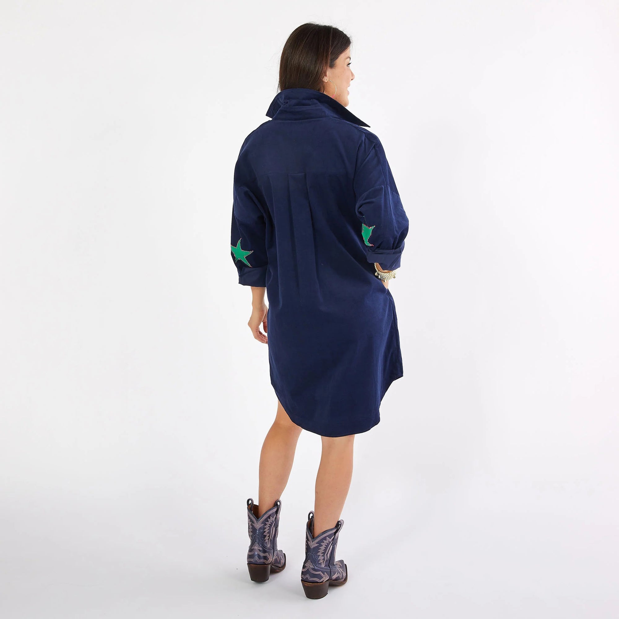 A person stands facing away, showcasing a contemporary chic style in the CARYN LAWN PREPPY Corduroy Dress. The dress is designed in a navy-blue hue and features hand-sewn green star patches on the sleeves. Their long hair cascades down to cowboy boots decorated with a star pattern, all set against a simple white studio backdrop that highlights the outfit's stunning details.