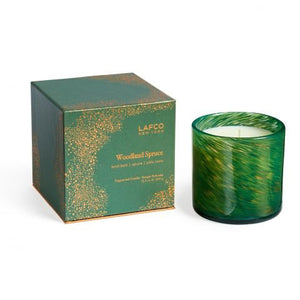 A lit soy wax candle with a green marbled design is positioned beside its coordinating box that reads "LAFCO - WOODLAND SPRUCE HOLIDAY CANDLE 15.5OZ." Housed in a hand-blown glass vessel, it emits the scent of a winter forest. The backdrop features a gradient of teal, enhancing the peaceful atmosphere.