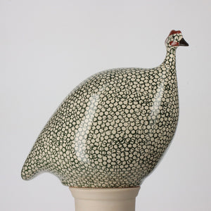 The LES CERAMIQUES DE LUSSAN - PETITE CERAMIC FRENCH GUINEA IN WHITE SPECKLED DUCK GREEN features a rounded body with black and white circles, a short neck, small head with a red patch, and pointed beak, elegantly perched on its white pedestal.