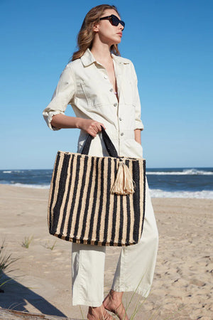 The MAR Y SOL - AMALFI TOTE BAG by MAR Y SOL is a yellow and white striped woven bag with two short handles and a large beige tassel near the top. It features a rectangular shape and textured fabric, offering a chic silhouette that exudes a handcrafted look.