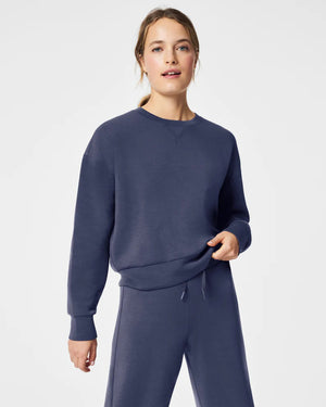 A woman stands confidently against a plain white background, wearing the SPANX Aireessentials Crew Neck Pullover and matching sweatpants made from luxurious comfort spacer fabric. She has her hair pulled back and her left hand is raised slightly. She smiles gently with her lips closed.