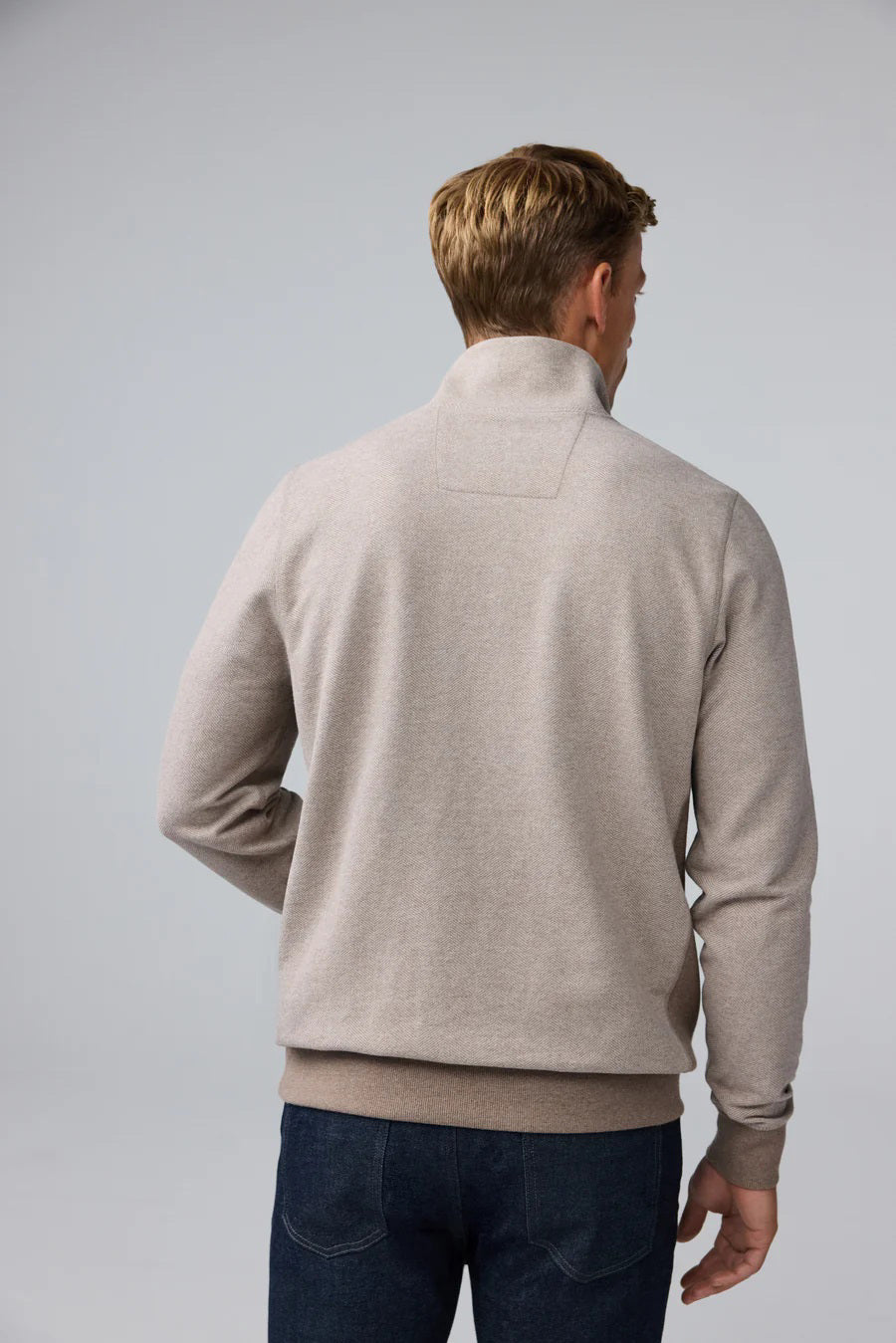 A man with short light brown hair stands in profile, wearing the MACK WELDON Ace 1/2 Zip Sweater in Herringbone and dark pants, one hand in his pocket, against a plain light gray background.