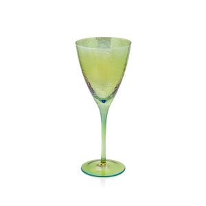 A round, golden tray features a selection of iridescent, textured glassware from the brand ZODAX, each with a capacity of 445 ml. This includes champagne flutes, wine glasses like the APERITIVO RED WINE GLASS - LUSTER GREEN, and tumblers. Two iridescent ornaments are artfully placed among the glassware, set against a dark, blurred background.