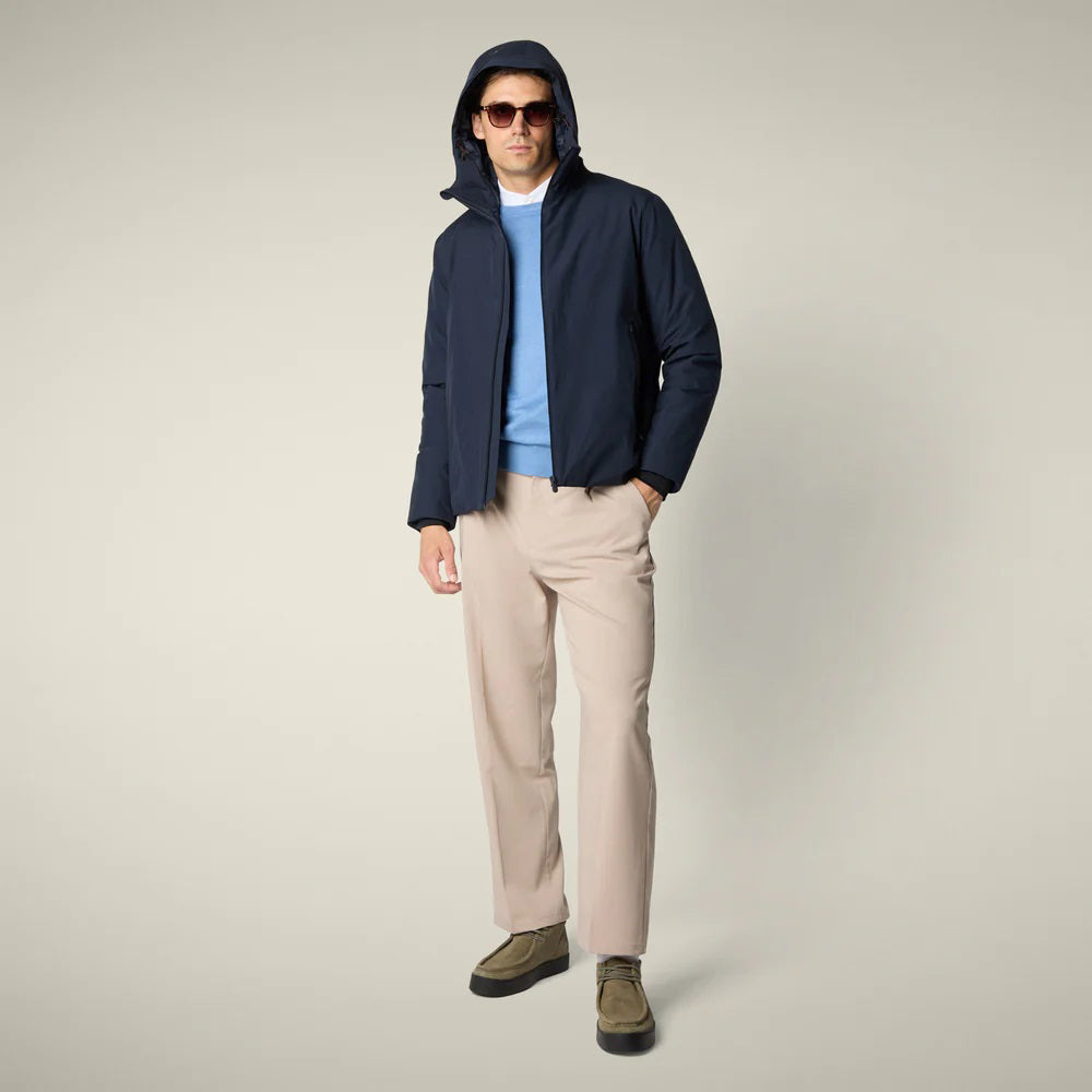A man in sunglasses, beige wide-leg pants, and olive green shoes wears the SAVE THE DUCK - FLYNN HOODED JACKET by SAVE THE DUCK. He stands against a light background facing forward, with one hand by his side and one near his zipper.