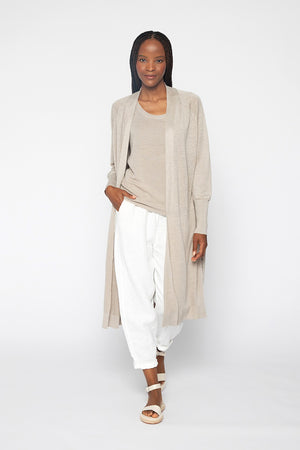 A person with long braided hair is wearing the KINROSS CASHMERE - LINEN DUSTER by KINROSS, a beige cardigan made from 100% linen, over a matching top and white loose pants. They are also wearing white sandals and standing against a plain white background. Their hands are in the duster's pockets, and they are looking straight ahead.