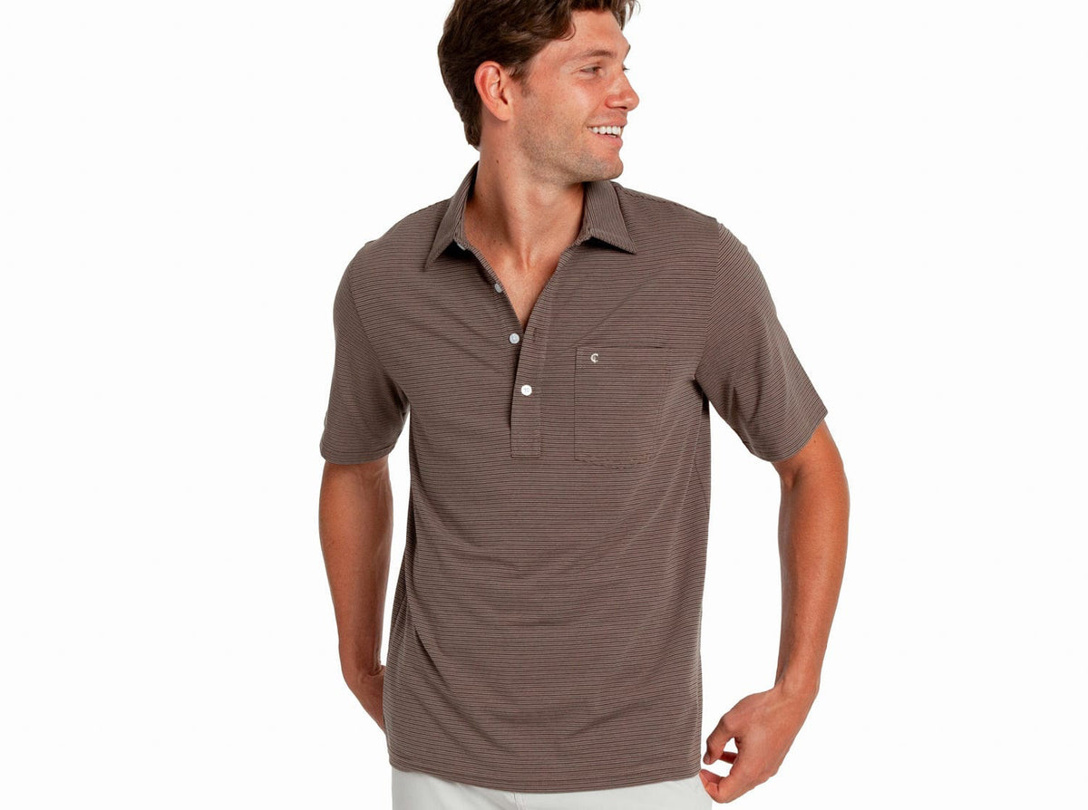 A man smiles and looks left, wearing the CRIQUET - PERFORMANCE PLAYERS SHIRT IN FERREL STRIPE TAUPE, featuring short sleeves, brown stripes, a chest pocket, and buttons, against a plain white background.
