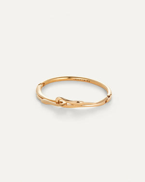 Wearing the JENNY BIRD Lisieux Bangle, designed like a horse bit with an elegant foldover clasp, a person rests their chin on clasped hands. They're dressed in a brown turtleneck sweater against a neutral background.