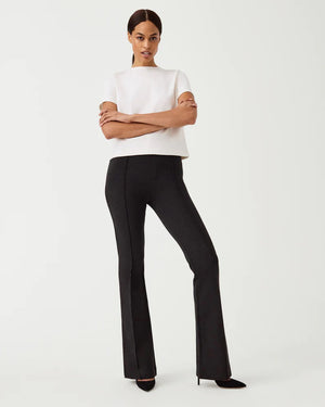 A person stands confidently against a plain background, wearing a white short-sleeved top and dark SPANX - Perfect Pant High Rise Flare pants made from smoothing premium ponte fabric. They have their arms crossed and are looking directly at the camera. They are also wearing black high-heeled shoes.