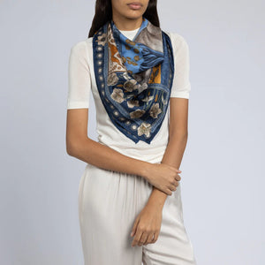 A person wearing a white short-sleeve shirt and light gray pants is shown with the SABINA SAVAGE - HONOURING ARGOS SILK TWILL SCARF 90CM by SABINA SAVAGE ENGLAND, featuring a blue and gold pattern reminiscent of Ancient Greek art, draped around their neck. The background is a plain light gray, and the person's hands are lightly clasped in front.