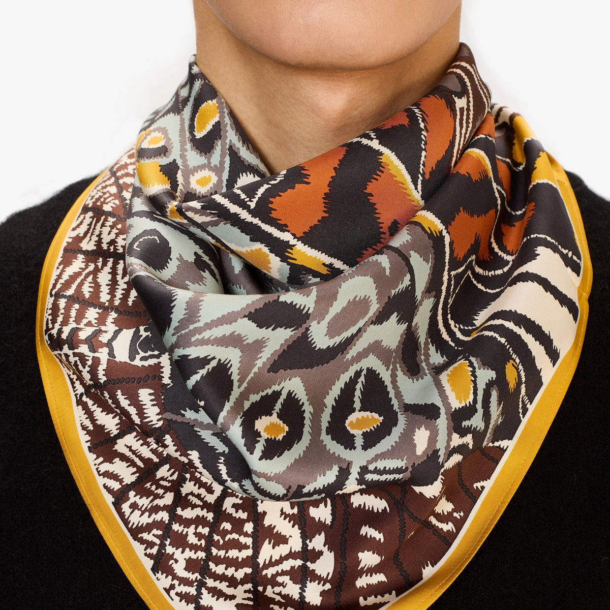 A close-up of a person wearing the INOUI EDITIONS - PAPILLON (BUTTERFLIES) SQUARE WOOL SCARF IN NATURAL by INOUI, draped around their neck over a black top. The scarf showcases abstract and geometric butterfly designs in earthy tones.