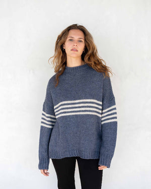 A person with long, wavy brown hair is wearing the MERSEA - SEACOAST STRIPE SWEATER, a loose-fitting, cozy blue knit sweater with white stripes on the sleeves, paired with black pants. They are standing against a plain, light-colored background and facing left, with their hands near their face.