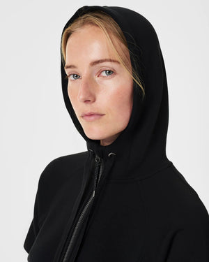 A person with blonde hair is wearing the SPANX AirEssentials Full Zip Hoodie made from ultra-soft fabric, with their hands in the pockets and a black skirt. They are standing against a plain, light-colored background and smiling softly.