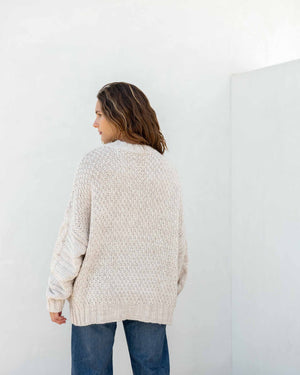 The image displays a detailed view of a person dressed in the MERSEA - TOPANGA CABLE SWEATER by MER SEA, showcasing its chunky knit cream-colored texture with large buttons on the sides, paired with blue jeans. The emphasis is on the sweater's intricate cable design and fine details, with the person's hand partially visible at their side.