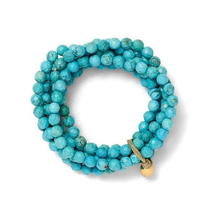 A person wearing a denim jacket showcases a stack of bracelets on their wrist, with the CC AND CO BY CATHERINE CANINO - SALLY BRACELET IN TURQUOISE standing out. Its stretchy fit bands and turquoise, white, and gold beads add natural color variations and texture to the accessory.