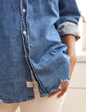 A person with curly hair stands in a kitchen, leaning back against a counter with hands resting on the surface. They wear an Eileen Relaxed Button Up Vintage Indigo Denim shirt from FRANK & EILEEN with rolled-up sleeves and white pants. The counter has a collection of wooden cutting boards and a vase with purple flowers.