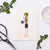 The PETAL PEOPLE PRESS - WITH SYMPATHY CARD by PETAL PEOPLE PRESS features a whimsical figure crafted from leaves and seeds, resembling a person holding a bouquet of Forget-Me-Not blue flowers. The head is fashioned from a green leaf, the body from a textured seed, and the legs and feet are thin twigs, making it perfect for adding delicate beauty to any sympathy card.