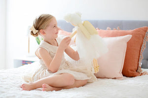 A young girl with light hair tied in a bow sits on a bed with white linens, smiling and holding up an Alimrose Ava Angel Doll Ivory Gold. She wears a light-colored dress, and behind her are pink and rust-colored pillows against a gray headboard.