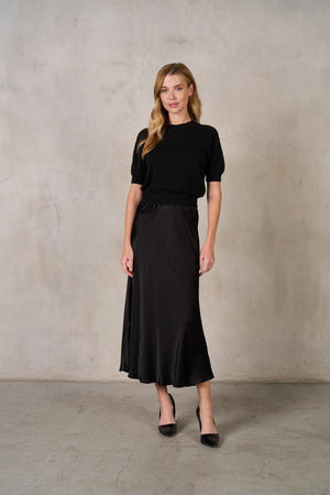 A person wearing a navy blue SATIN MIDI SKIRT from ROSE & CROWN paired with a matching navy blue top. The individual is also wearing beige pointed-toe heels. The background features a plain, light-colored wall with a concrete floor.