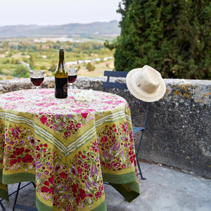 A round outdoor table with a COULEUR NATURE JARDIN RED AND GREEN TABLECLOTH 71X71, featuring floral designs. A fruit tart sits at the center, surrounded by rustic metal chairs akin to Southern France gardens, with lush greenery providing a picturesque backdrop.