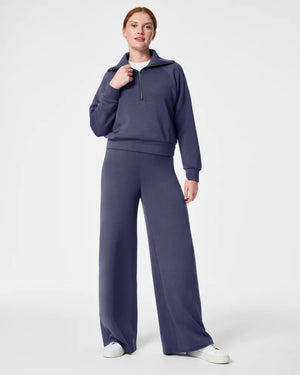 A person stands against a plain background wearing a matching two-piece outfit. The ultimate throw-on-and-go style consists of a long-sleeve, quarter-zip, high-collar pullover and SPANX AIRESSENTIALS WIDE LEG PANT in a deep blue color. They also wear white sneakers and have their hair pulled back.