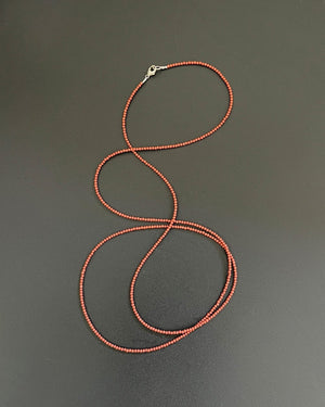 The ANN LIGHTFOOT - RED JASPER STRAND NECKLACE by ANN LIGHTFOOT, featuring small, round dark red beads, is laid out on a white surface with its 39-inch length forming loose curves. This delicate piece is fastened with a sterling silver clasp.